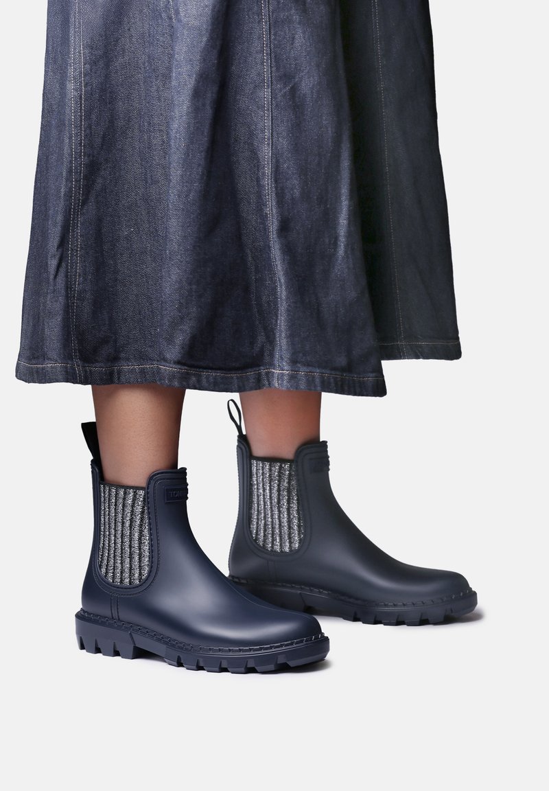 Personne portant des bottines de pluie bleu foncé avec des panneaux en tissu extensible gris et une jupe en jean foncé.