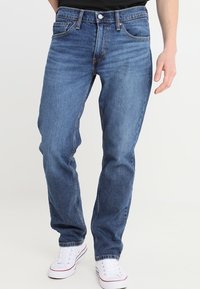 Man med klassiska blå raka jeans och vita sneakers, stående mot en enkel ljus bakgrund.