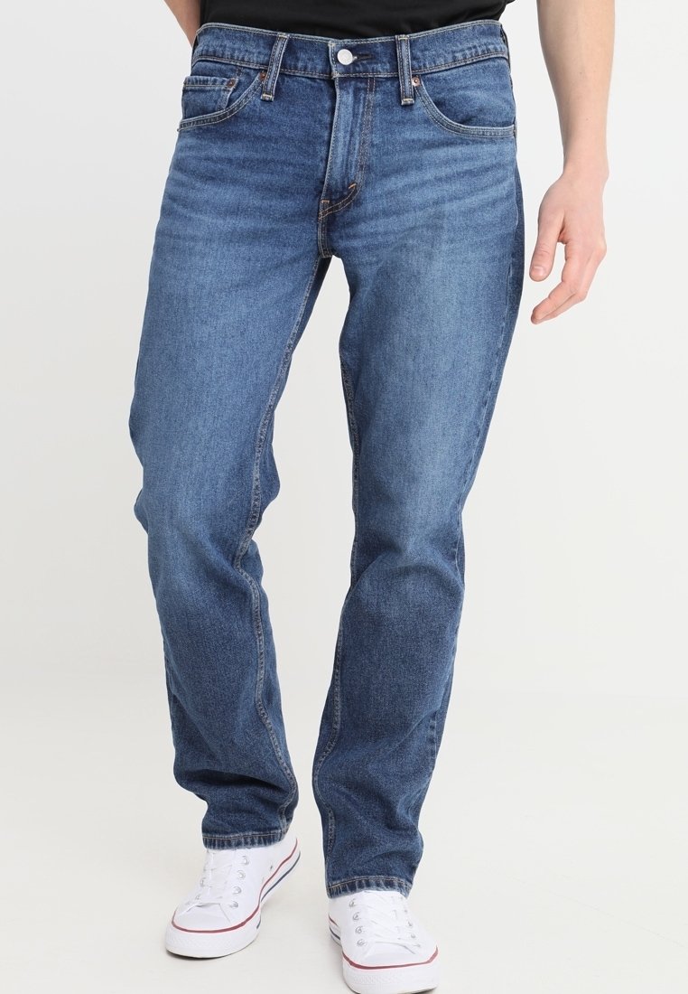 Man med klassiska blå raka jeans och vita sneakers, stående mot en enkel ljus bakgrund.