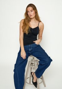 Vrouw met lang haar die op een krukje zit, draagt een zwart hemdje, wijde blauwe jeans en zwarte puntige schoenen tegen een witte achtergrond.