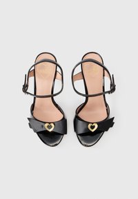 Black patent leather high-heeled sandals featuring a bow detail with a gold heart accent and adjustable ankle strap. Beige insole with logo.