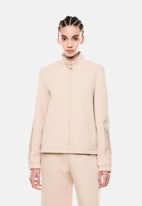 Felpa zip con collo alto di un beige chiaro, maniche testurizzate con delicati abbellimenti e una vestibilità morbida.