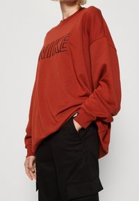 Person klädd i en lös, röd Nike-tröja med svarta cargobyxor, en hand i fickan, mot en ljus, enfärgad bakgrund.