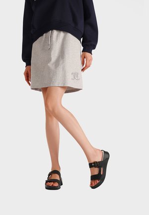 Persona che indossa una gonna grigia con coulisse, un piccolo logo e sandali neri con fibbia, in piedi con una gamba incrociata davanti all'altra.