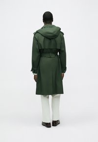 Trench verde scuro con cappuccio, vita con cintura e maniche lunghe, visto da dietro, abbinato a pantaloni chiari e scarpe scure.