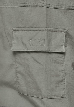 Tessuto grigio con una trama liscia presenta una tasca rettangolare con una patta, delineata da cuciture in contrasto. Design semplice con dettagli minimi.