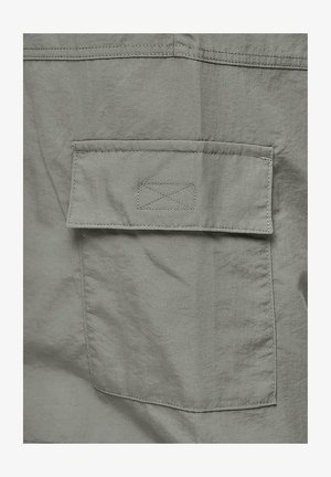 Szara tkanina o gładkiej fakturze z prostokątną kieszenią z klapką, wykończoną kontrastowym szwem. Prosty design z minimalną ilością detali.