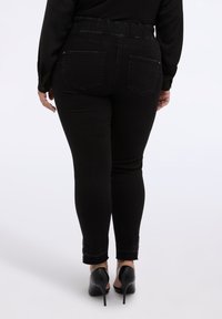 Jeans skinny de mezclilla negra con cintura alta y banda elástica, que cuentan con dos bolsillos traseros y detalles decorativos en el dobladillo, combinados con tacones negros.
