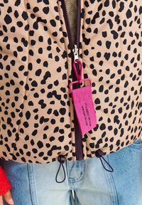 Puchowa kurtka w jasnym beżu z czarnymi plamkami lamparta, z fioletowym zamkiem błyskawicznym i jasnoróżową metką. Regulowane wykończenie z sznurkami.