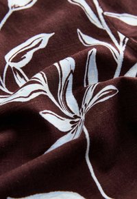 Bordeauxroter Stoff mit strukturierter Oberfläche, der weiße Blumenmuster zeigt. Das Design umfasst filigrane Blütenblattformen und abstrakte Linien.