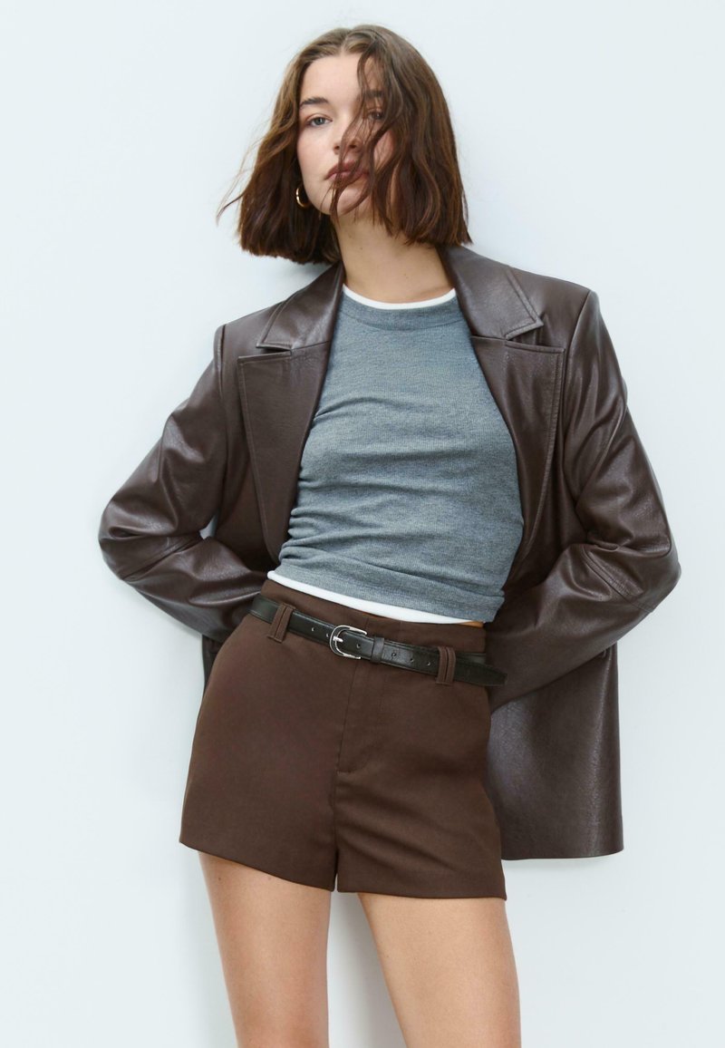 Donna con capelli castani fino alle spalle che indossa orecchini a cerchio dorati, giacca di pelle marrone, top corto grigio e pantaloni corti marrone con cintura.