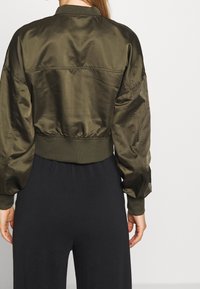 Kakijsově zelená bomber bunda s lesklou, hladkou texturou, zkráceným designem, žebrovanými manžetami a přiléhavým lemem, nošená přes černé kalhoty.