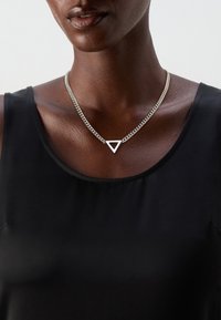Collier en chaîne argentée avec un pendentif triangulaire, présentant une surface lisse, suspendu près de la clavicule, porté sur un haut sans manches noir.