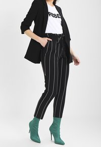 Černý blazer přes bílý tričko s textem "REBEL", doplněný černo-bílými pruhovanými kalhotami a zelenými špičatými kotníkovými botami.