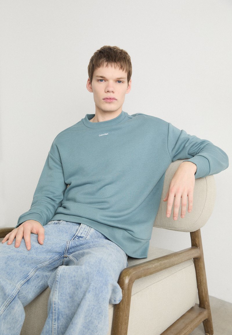 Sudadera azul claro con cuello redondo y pequeño logotipo de Calvin Klein, combinada con jeans de mezclilla claros, sentado en una silla tapizada beige.