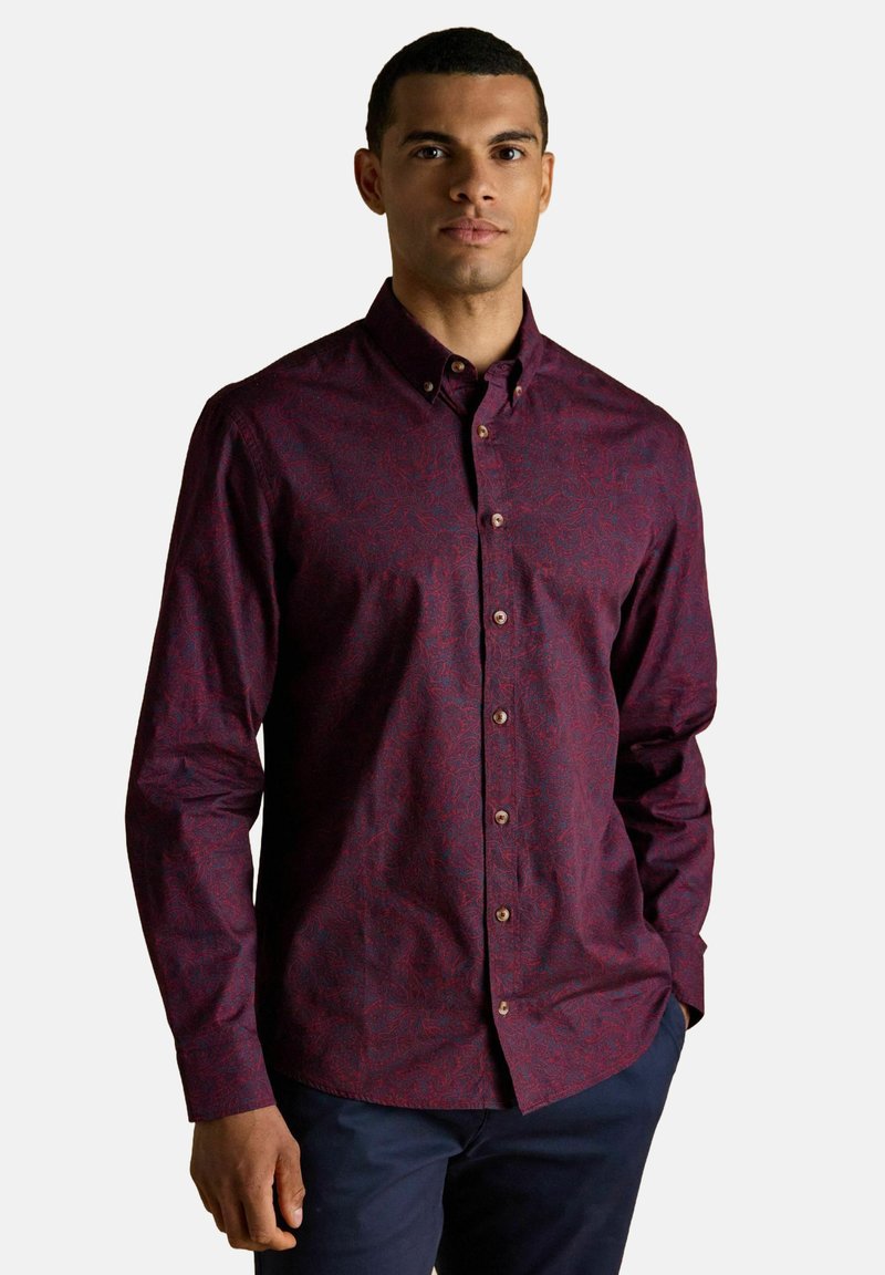 Vīrietis, kas uzvilcis tumši sarkanu, rakstainu krekla ar garām piedurknēm, stāv ar rokām tumši zilās biksēs kabatās uz vienkārša fona.