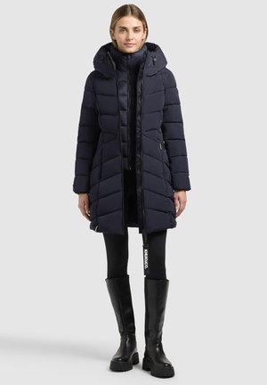 Femme portant un long manteau matelassé bleu marine avec capuche, des leggings noirs et des bottes noires épaisses montantes, debout devant un fond uni.