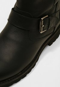 Bottines en cuir noir avec un petit talon, présentant une sangle décorative à boucle et une texture en relief. Semelle en caoutchouc robuste avec un motif de traction.