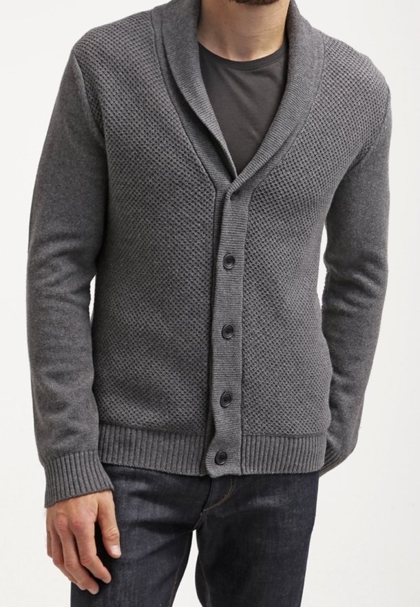 Homme portant un cardigan boutonné texturé gris foncé par-dessus un t-shirt noir avec un jean foncé, debout devant un fond blanc.