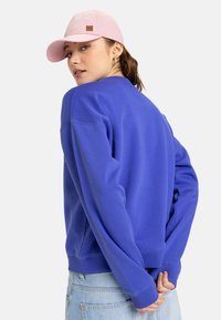 Sudadera de algodón morada con un corte relajado, puños y dobladillo de canalé; combinada con una gorra de béisbol rosa claro que presenta un parche con un logo.