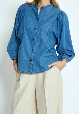 Femme portant une chemise en denim bleue à manches longues bouffantes boutonnée, associée à un pantalon large beige clair, les mains dans les poches.