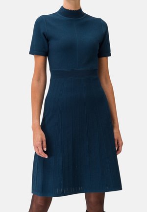 Granatowa sukienka dzianinowa z krótkim rękawem, dopasowana w talii, z teksturowanymi pionowymi wzorami na spódnicy, noszona przez stojącą osobę.