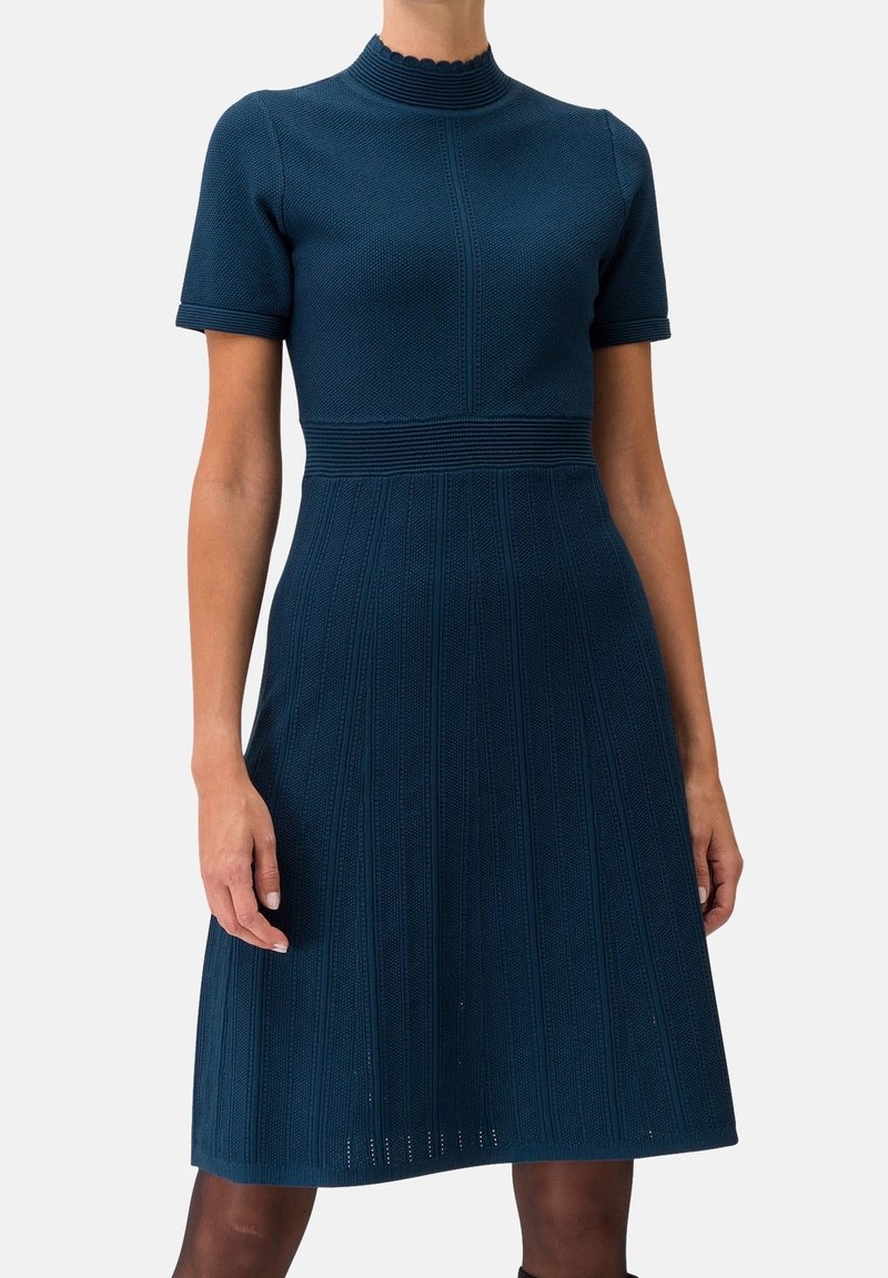 Robe en maille bleu marine à manches courtes avec taille cintrée et motifs verticaux texturés sur la jupe, portée par une personne debout.