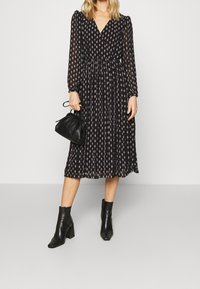 Zwart midi-jurk met een V-hals en lange pofmouwen, voorzien van een bloemenpatroon, vloeiende stof en een geraffineerde taille. Gecombineerd met zwarte enkellaarsjes en een kleine tas.