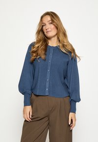 Blauwe geribbelde cardigan met een ronde halslijn, knoopsluiting en ballonmouwen, gecombineerd met bruine wijde broekspijpen.