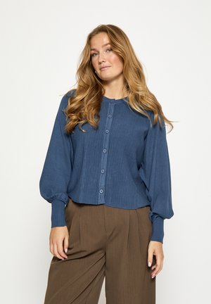 Modrý rebrovaný kardigán s okrúhlym výstrihom, zapínaním na gombíky a balónovými rukávmi, skombinovaný s hnedými nohavicami širokého strihu.