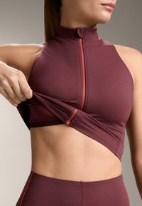 Bordó atlétikai felső magas nyakkal és cipzáras záródással, narancssárga díszítésekkel és testhezálló kialakítással, viselés közben igazítva bemutatva.