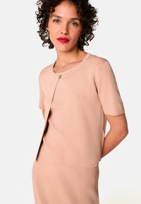 Kurzärmliges, hellrosa Oberteil mit rundem Ausschnitt und einem kleinen goldenen Hardware-Detail an der Vorderseite, gefertigt aus einem strukturierten Stoff.