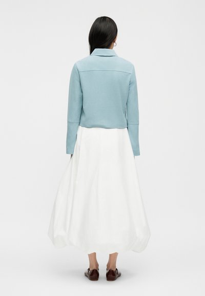 Γυναίκα με μακριά σκούρα μαλλιά, που φοράει ανοιχτό μπλε μπουφάν, λευκή φούστα μέχρι τον αστράγαλο και καφέ παπούτσια, στέκεται με την πλάτη γυρισμένη σε απλό φόντο.