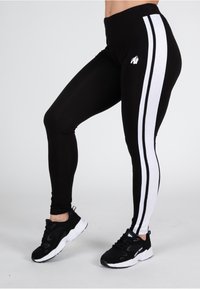 Leggings neri con strisce laterali bianche, con un logo sulla coscia. Realizzati in materiale elasticizzato, progettati per l'abbigliamento attivo. Abbinati a sneaker nere.