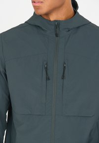 Dunkelgrüne, leichte Kapuzenjacke mit einem Frontreißverschluss, zwei Brusttaschen und verstellbaren Kordelzügen an der Kapuze und den Taschen.