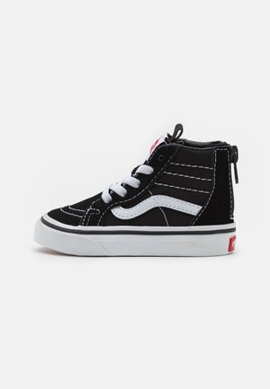 Sort-hvid high-top sneaker med hvide snørebånd, sideliste, syningsdetaljer og en rød logo på hælen mod en ensfarvet baggrund.