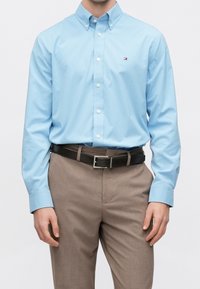 Homme portant une chemise bleu clair à boutons rentrée dans un pantalon beige avec une ceinture noire sur un fond uni.