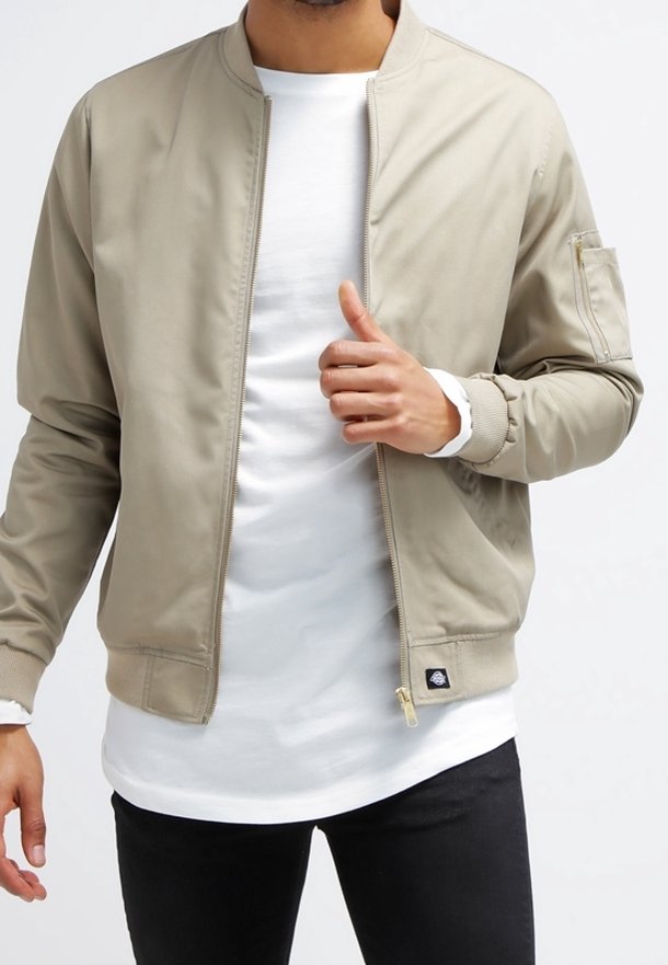 Homme portant une veste bomber beige par-dessus un t-shirt blanc et un pantalon noir, tenant la fermeture éclair de la veste près de la poitrine.