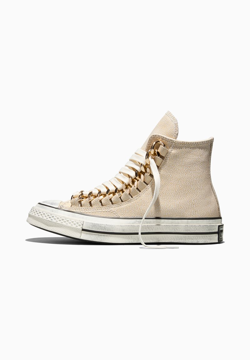 Beige high-top sneaker with white sole and laces threaded through large gold metal eyelets on a white background.