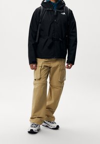Veste imperméable noire avec taille ajustable, associée à un pantalon cargo beige doté de multiples poches. Des baskets athlétiques noires et blanches complètent le look.