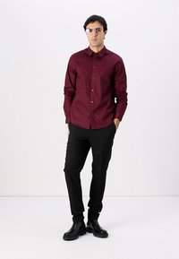Bordeauxfarbendes Hemd mit langen Ärmeln, schwarze Hose und schwarze Knöchelstiefel. Zu den Accessoires gehören eine reflektierende Brille. Einfaches, maßgeschneidertes Design.