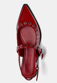 Rote Lacklederschuhe mit spitz zulaufender Zehe, ausgestattet mit kreisförmigen silbernen Ösen und einem Knöchelriemen mit Schnalle. Texturiertes Fußbett.