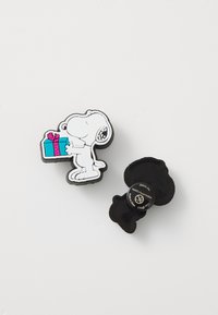Charm en forme de Snoopy avec un cadeau, présentant un corps blanc et des accents colorés. Dos noir, texture lisse, conçu pour être attaché à des chaussures ou des accessoires.