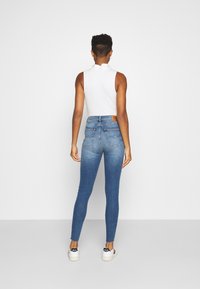 Jeans ajustados en denim azul oscuro con un lavado desgastado, cintura alta, diseño de cinco bolsillos y un parche de cuero en la parte trasera. Combinados con una blusa blanca sin mangas.