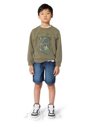 Junge trägt ein olivgrünes Sweatshirt mit Schlangen- und Gitarrendesign, blaue Jeansshorts und schwarz-weiße Sneakers mit geöffneten Schnürsenkeln.