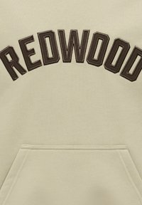 Sweatshirt beige avec des lettres en cuir marron foncé cousues formant le mot "REDWOOD" en arc au-dessus d'une poche kangourou avant.