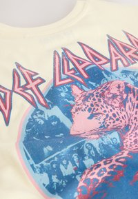 Maglietta di cotone giallo chiaro con una grande grafica che presenta un leopardo rosa su uno sfondo blu, sovrapposta a immagini vintage di concerti.