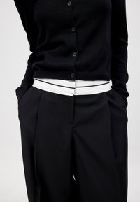 Schwarze Strickjacke mit Knopfverschlüssen, kombiniert mit hochtaillierten schwarzen Hosen, die einen weißen Gürtel und ein flaches Vorderdesign aufweisen.