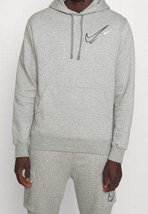 Mann trägt hellgrauen Hoodie und passende Sweatshorts, beide mit schwarzen und weißen Nike-Swoosh-Logos auf der linken Brust und dem linken Oberschenkel.