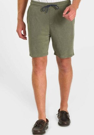 Man draagt olijfgroene short met trekkoord en bruine leren casual schoenen, staand tegen een effen witte achtergrond.